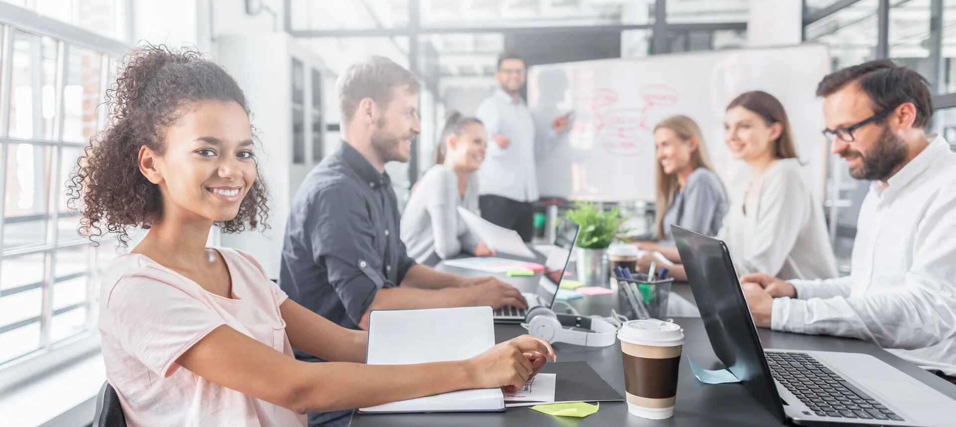 jeune femme qui regarde l'objectif en souriant avec 6 collègues en open space en arrière plan