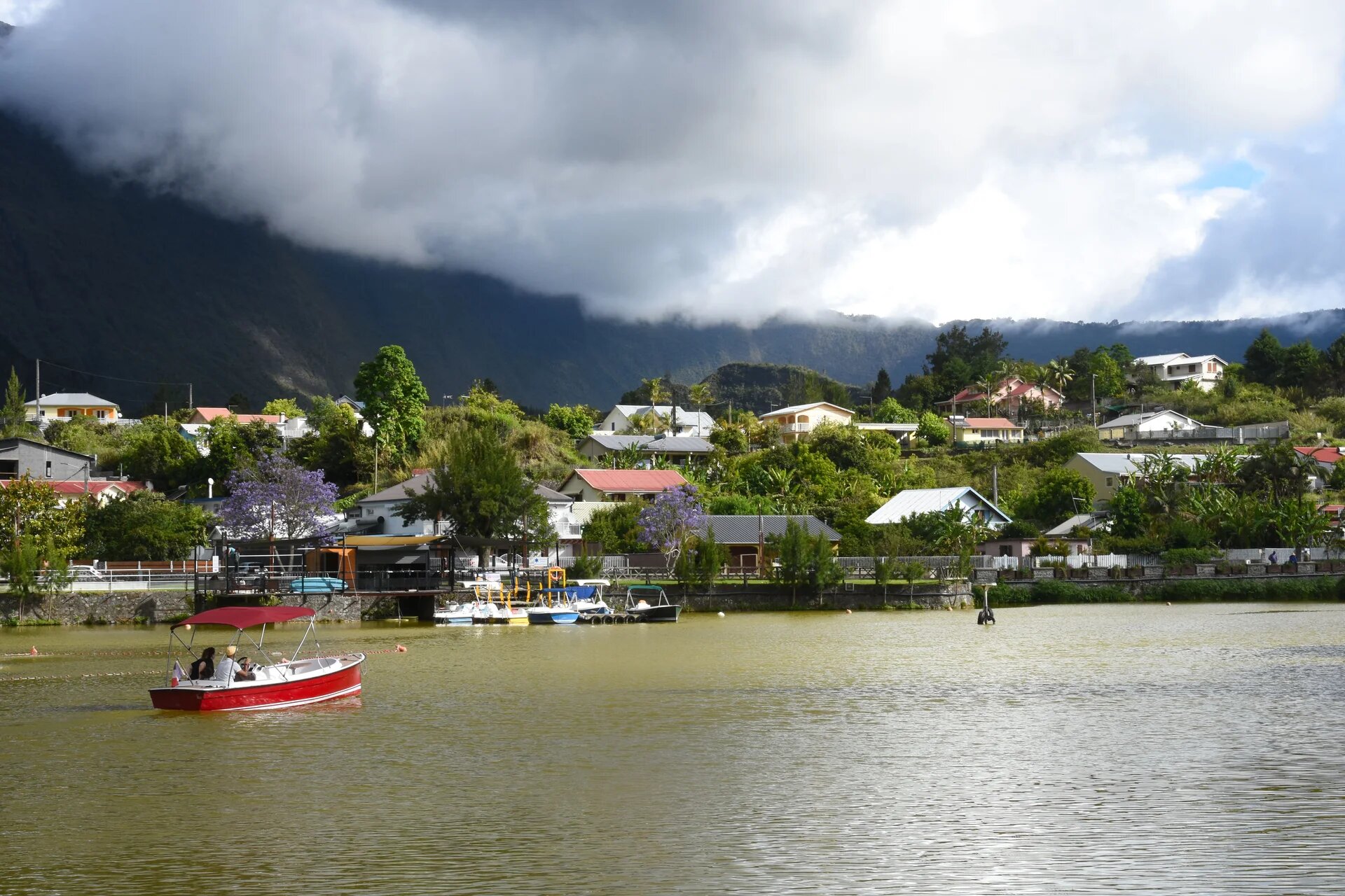 Vue d'un bord de mer avec de la végétation et des maisons. au premier plan un petit bateau de plaisance et en arrière plan la montagne et les nuages