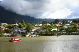 Vue d'un bord de mer avec de la végétation et des maisons. au premier plan un petit bateau de plaisance et en arrière plan la montagne et les nuages